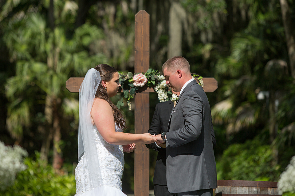 bride and groom exchange wedding rings in front of a wooden cross