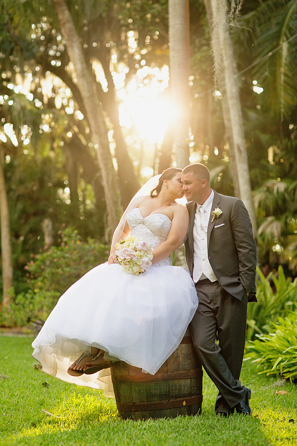bride sitting on a barrell kissing her husbands check with the sunlight peaking through the trees