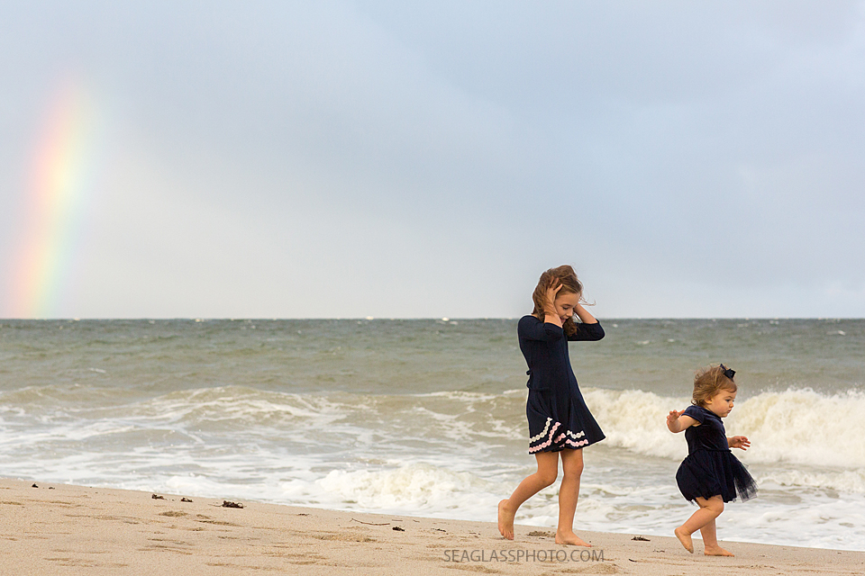 sisters wearing navy dresses walking on the shoreline away from the rainbow in Vero Beach Florida family photography