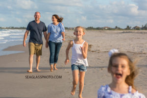 Two daughters running toward the photographer while parents walk behind on the beach for family photo shoot in Vero Beach Florida