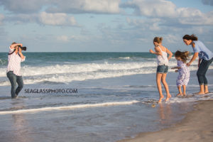 Photographer shooting in the water taking pictures of a mom and her two daughters jumping over the waves on the beach during family photo shoot in Vero Beach Florida