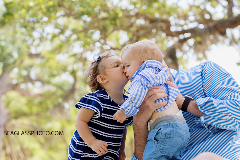 big sister kissing her little brothers head while father holds him during family photos at Riverside park in Vero beach Florida