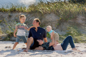 Family sit together on the beach with mom holding her youngest son and the father laughing with their oldest son on the beach during family photo shoot in Vero Beach Florida