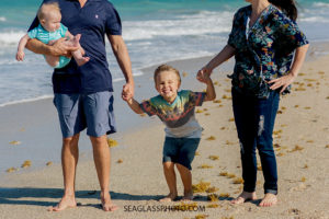 Close up of the oldest son holding mom and dads hands and jumping up and down on the beach while his younger brother looks at him during family photos in Vero Beach Florida