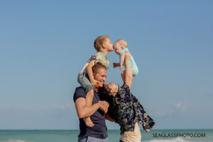Mom and dad holding their sons in the air while big brother kisses his little brother on his forehead during family photos on the beach in Vero Beach Florida