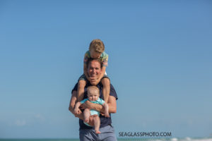 Dad holding his two sons, oldest on his shoulders and youngest in his arms while oldest son kisses his head during family photos on the beach in Vero Beach florida