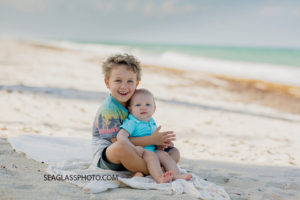 Big brother holds is little brother while sitting on the beach with the ocean behind them during family photos in Vero beach Florida