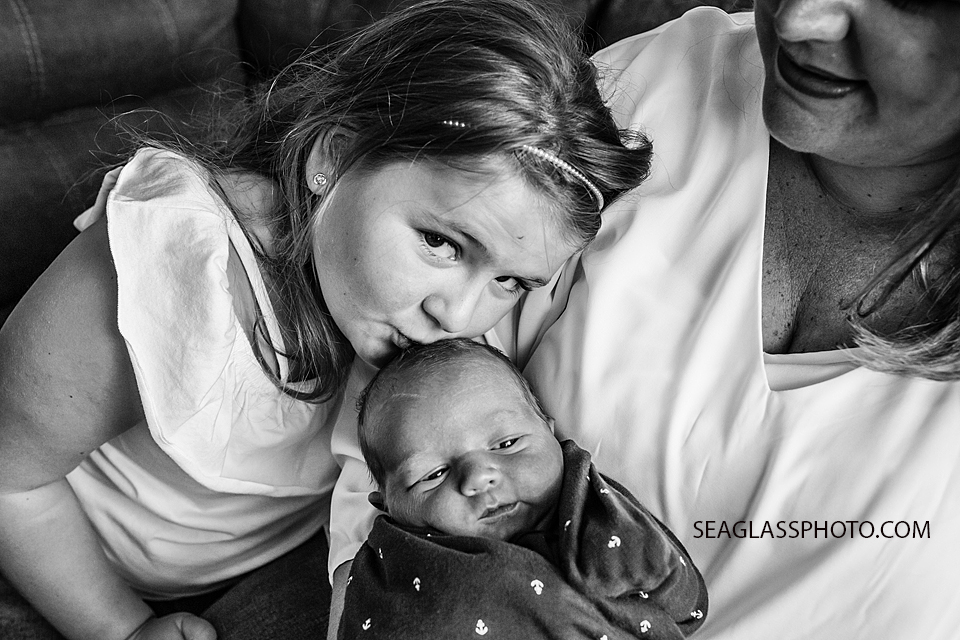 Black and white photo of oldest sister giving her little brother a kiss on the forehead as mom holds him during newborn session in Vero Beach Florida