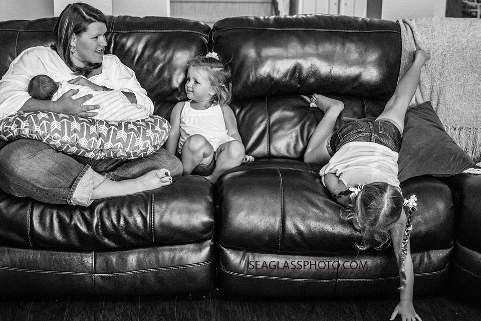 Black and white photo of mom feeding her newborn so talking to her youngest daughter as her middle daughtger messes around on the couch during newborn session in Vero Beach Florida