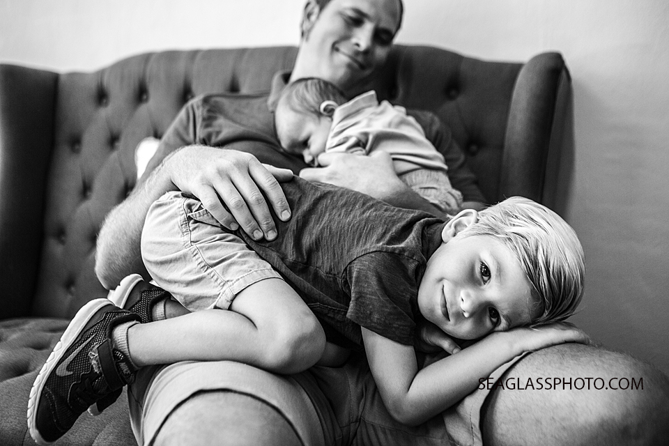 Black and white photo of young boy laying on his fathers lap with his little brother above him during newborn session in Downtown Vero Beach Florida