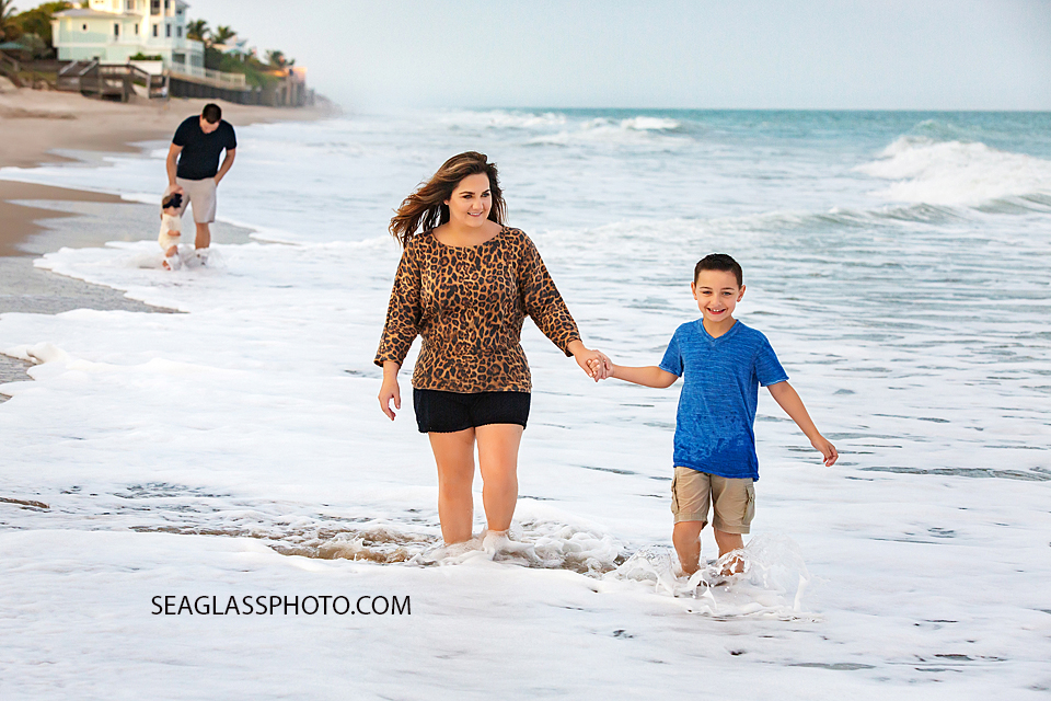 Mother holds hands with her son walking on the beach with the dad and daughter following behind during their family photoshoot in Vero Beach Florida