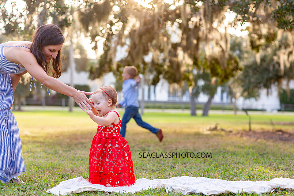 mother helps her daughter stand up while big brother plays in the back ground during family photos at Riverside in Vero Beach Florida