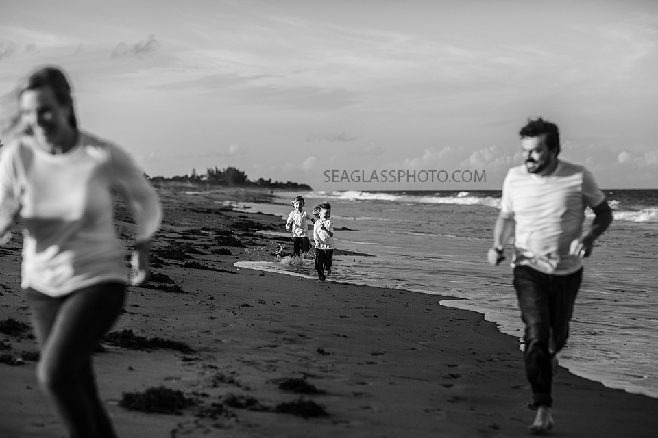 Black and white photo of two boys running along the water behind their parents during family photoshoot in Vero Beach Florida