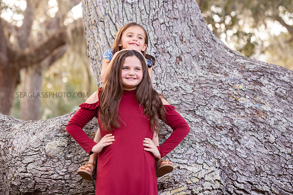 younger sister smiles behind her big sister while sitting on a tree during sister photoshoot in Vero Beach Florida
