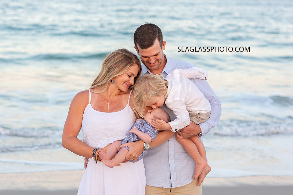 Big brother kisses his little brothers forehead while the parents hold them on the beach during their family photoshoot in Vero Beach Florida