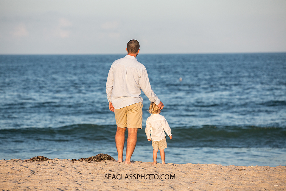 Father looking out onto the ocean with his oldest son on the beach during their family photoshoot in Vero Beach Florida