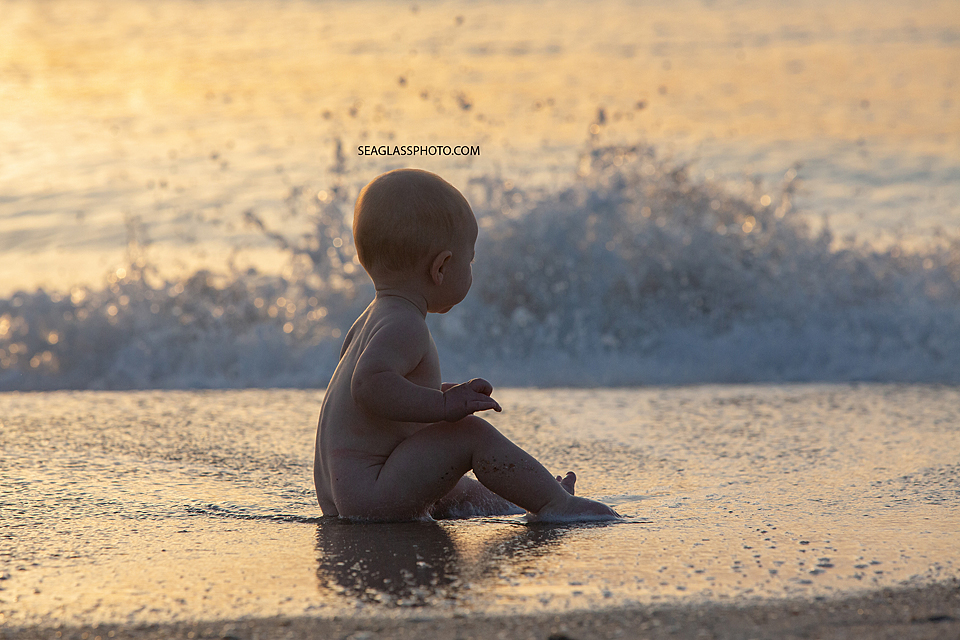 Naked baby sitting in the sand as waves roll up to him on the beach at sunrise in Vero Beach Florida