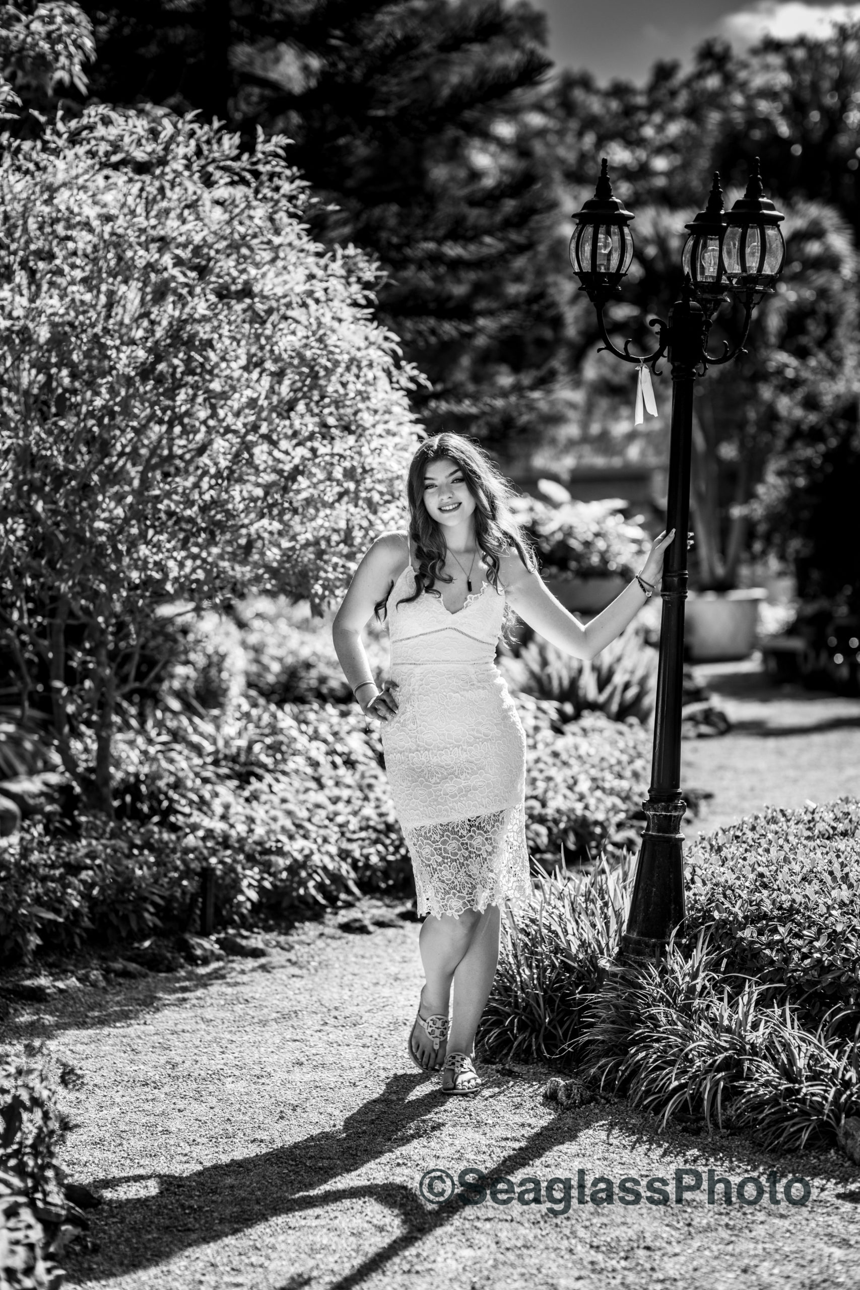 black and white portrait of high school senior girl with long brown hair and wearing a white lace dress surrounded by flowers at Rocky City Gardens in Vero Beach FL
