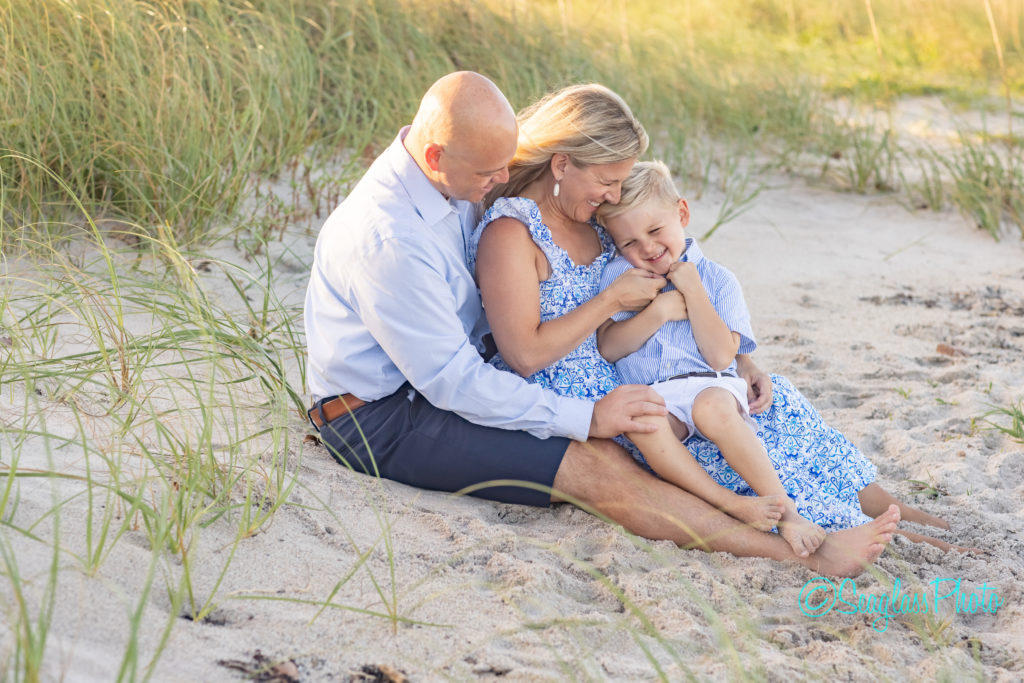 family of 3 sitting in the sand dunes tickingl each other laughing. Moorings club in Vero Beach 