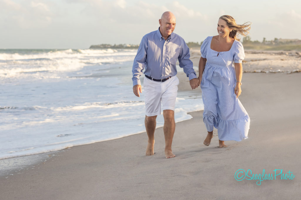 couple walking down the beach wearing blue and white. The wind blowing in her hair and they both have big smiles 