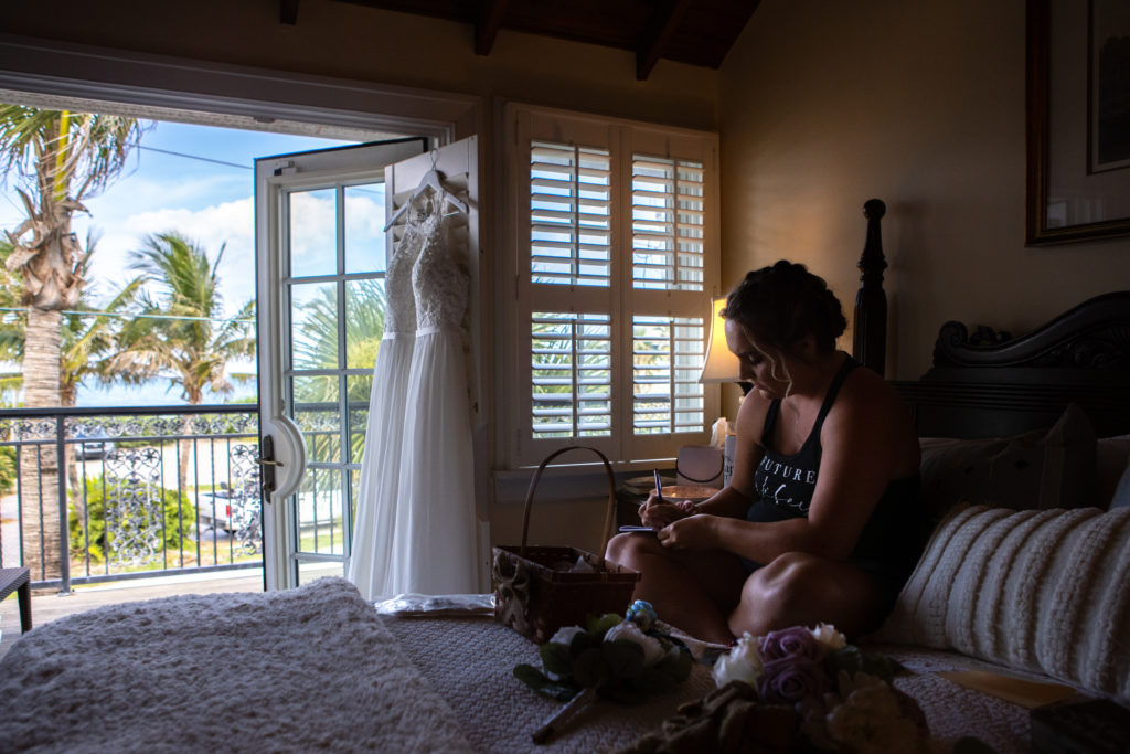 Bride sitting on the bed at Port d'Hiver Bed and Breakfast with her wedding gown hanging on the door overlooking the ocean