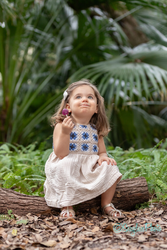 toddler girl sitting on a log surrounded by ferns and palms while holding a purple lolipop