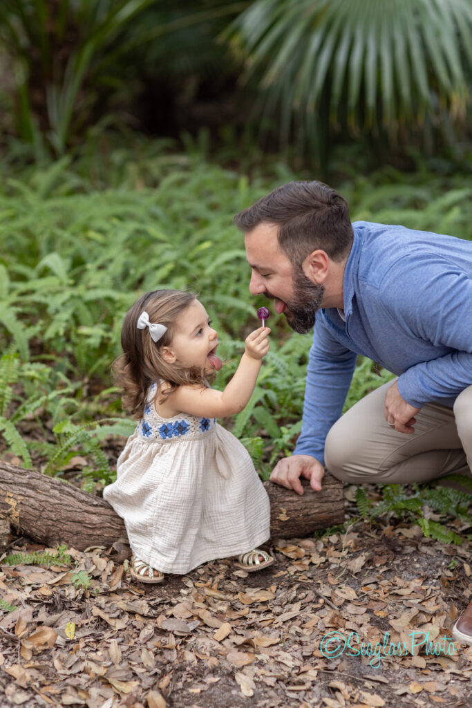 toddler girl giving her dad a purple lolipop while sitting on a log surrounded by ferns in Vero Beach Florida