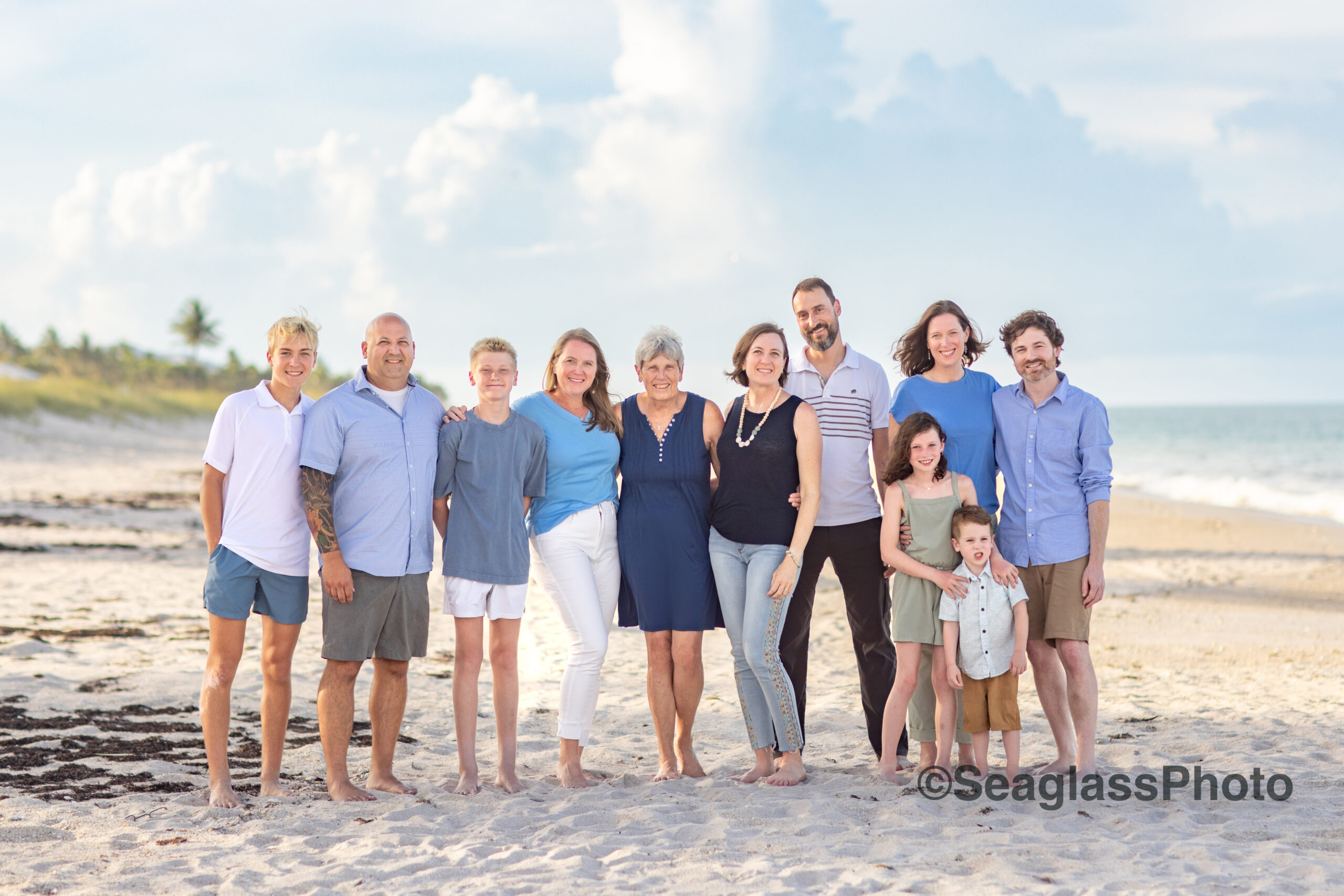 grandma with her adult kids and grandkids on the beach wearing an array of blues and white with a pretty sky behind them