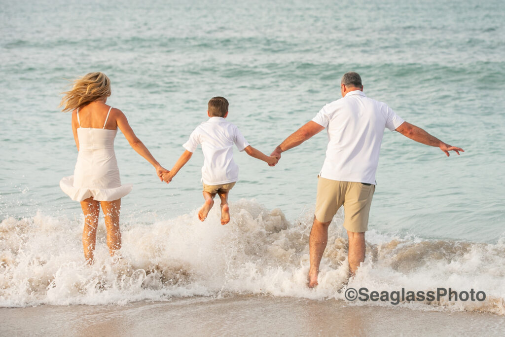 parents holding their sons hands jumping in the waves family portrait in Vero Beach Florida wearing a mix of white, khaki and cream