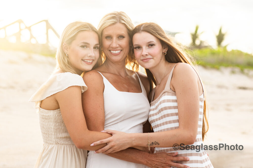 stunning portrait of mother and two teen daughters hugging on the beach with sunlight shining 