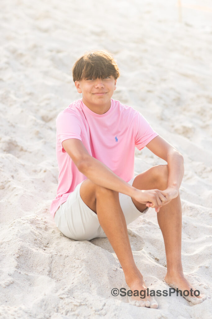 teen boy sitting on the beach wearing a pink shirt and khaki shorts with a silver cross necklace at the Disney Vero Beach Resort