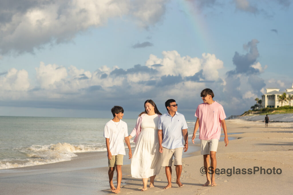 mom and dad walking on the beach with their two sons wear white, pink and blue. A rainbow is shining onto the ocean in front of the Disney Vero Beach Resort 