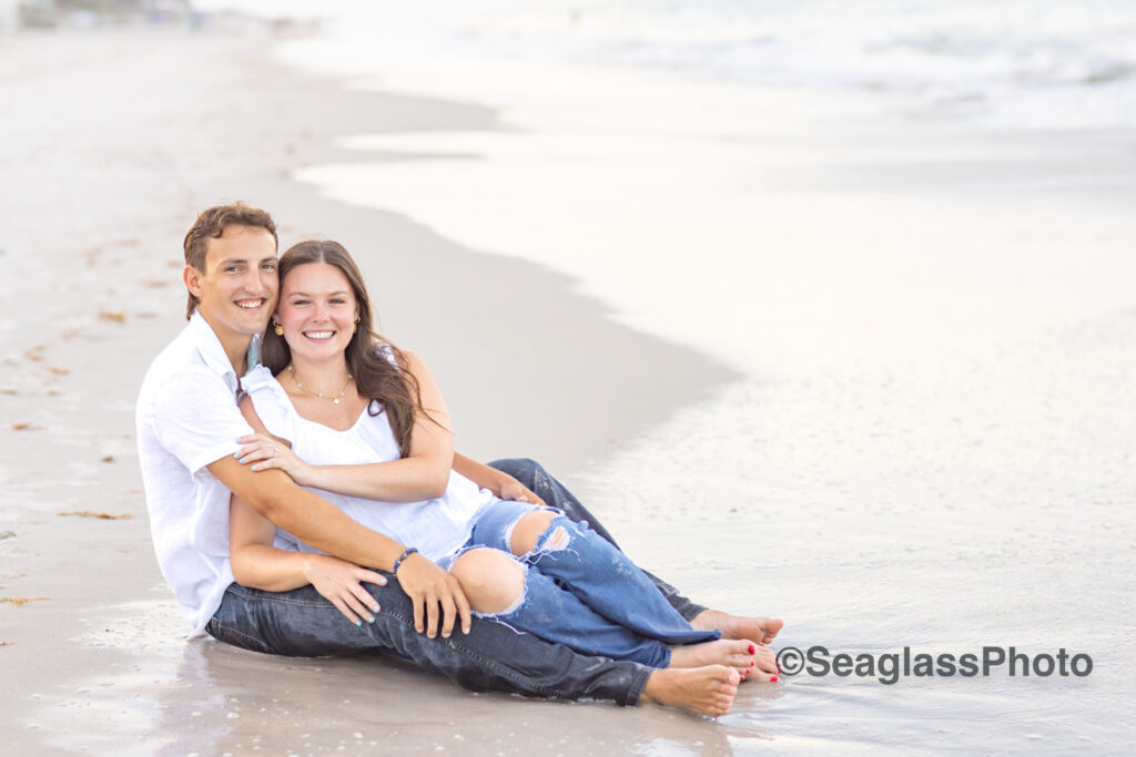 couple wearing white shirts and jeans sitting on the  beach in Vero Beach Florida engagement photoshoot