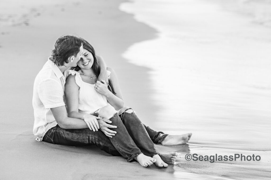 black and white photo couple wearing white shirts and jeans sitting on the  beach in Vero Beach Florida engagement photoshoot