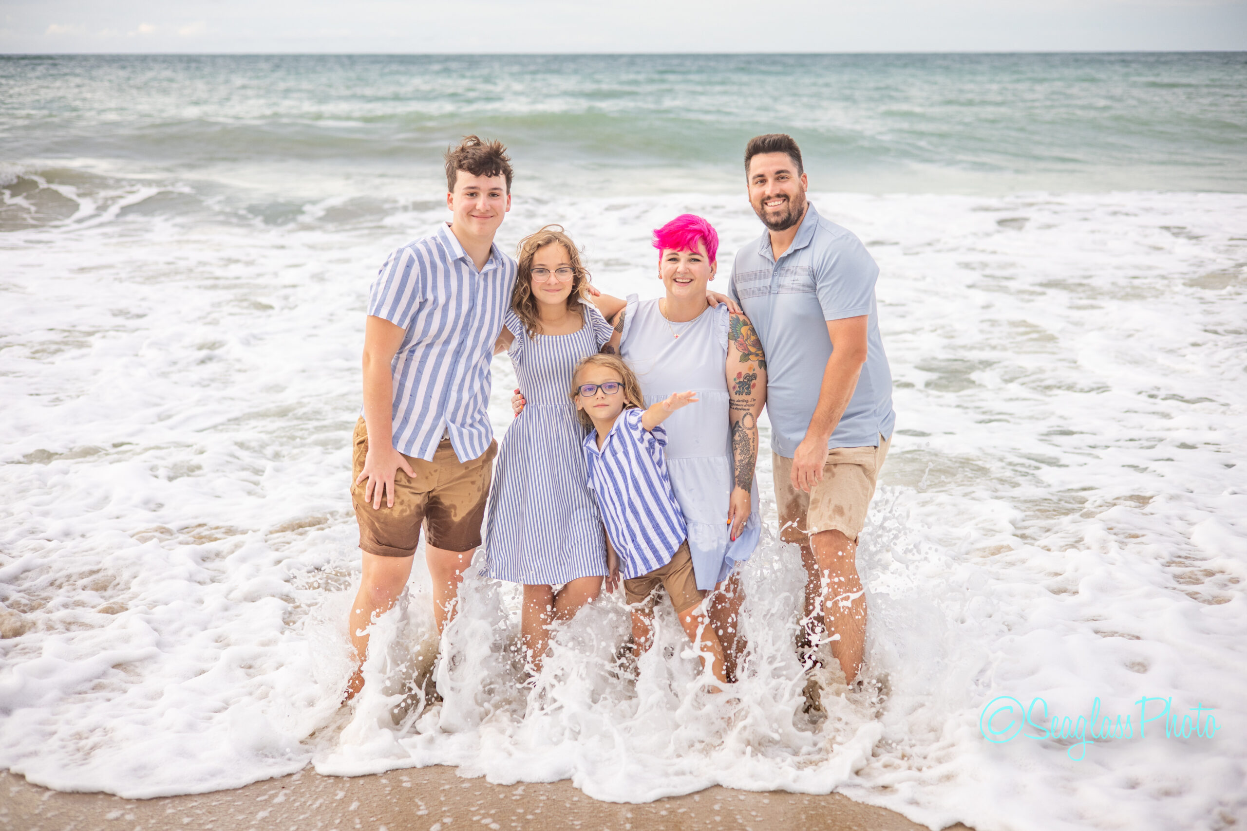 A family of five, smiling and standing in shallow ocean waves at the beach, enjoy a Disney DVC vacation. They wear coordinated blue and white outfits—one with bright pink hair—under a clear sky above the shimmering sea. Vero Beach Photoshoot