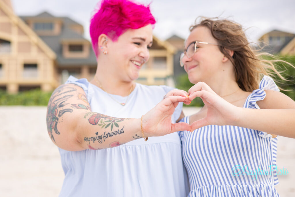 Two women stand close together on a beach, smiling and forming a heart shape with their hands—one with bright pink hair and tattoos, the other with long light brown hair and glasses. Disney DVC fans might spot familiar resort buildings in the background. Vero Beach Photoshoot