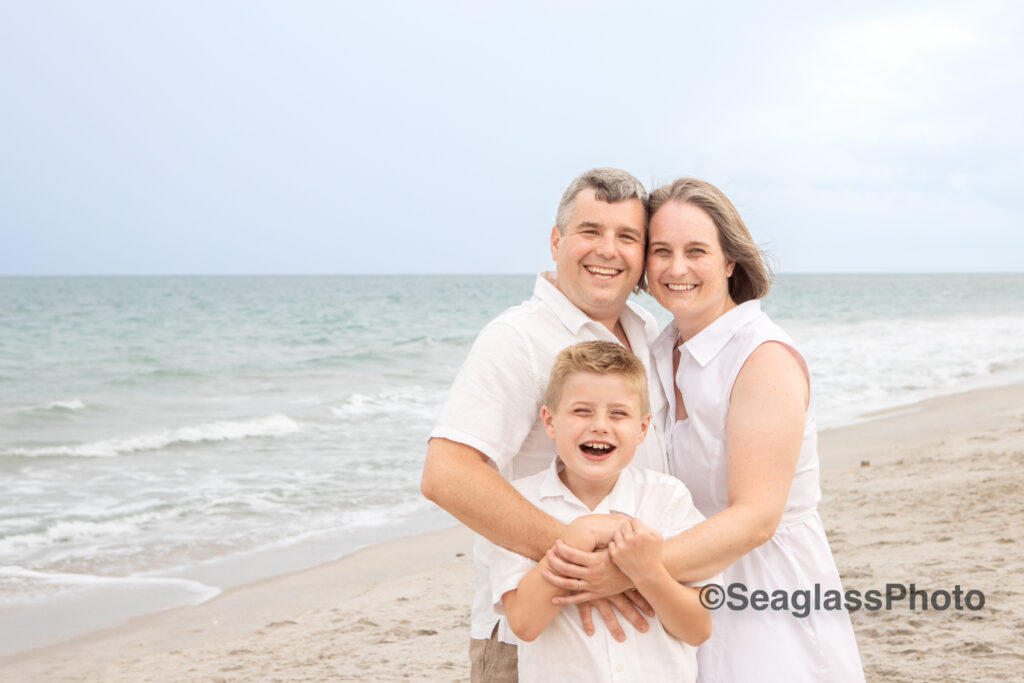 Family photo of mom and dad hugging onto their son wearing all white on the beach in Florida 