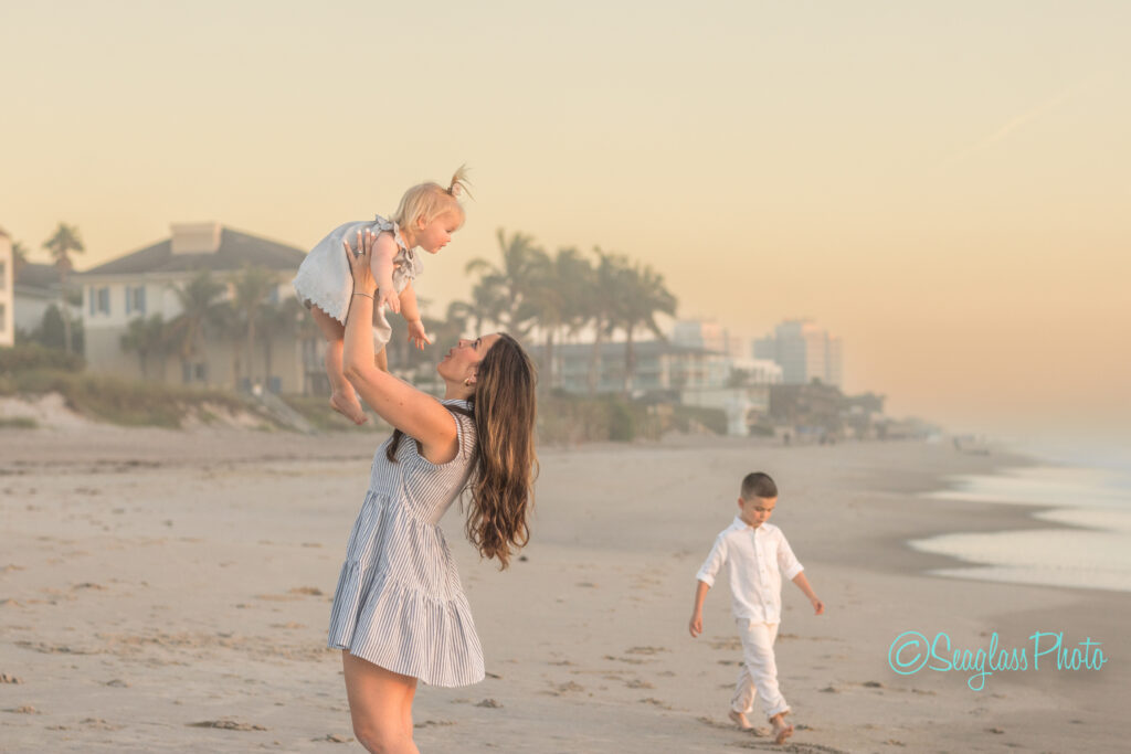 A woman in a striped dress lifts a smiling toddler in the air on a sandy beach at sunset. A young boy in white walks nearby. Palm trees and beach houses are in the background. Vero Beach Photoshoot