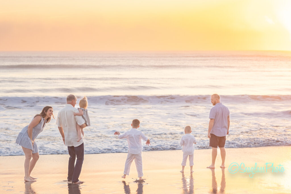 A family of five, including two adults and three children, enjoy a sandy beach at sunset. The adults are smiling and interacting with the children, who are looking at the waves. The ocean and colorful sky create a serene backdrop. Vero Beach Photoshoot