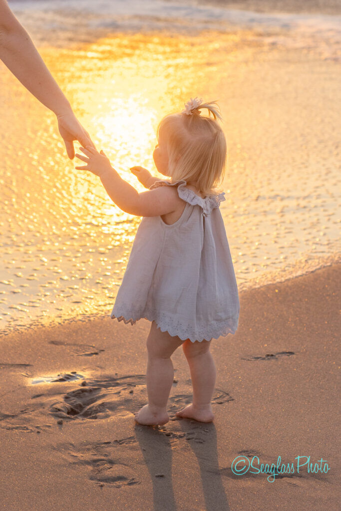A toddler in a light dress stands on the beach, reaching towards an adults hand. The sun sets over the water, casting a golden glow on the scene. Footprints are visible in the wet sand. Vero Beach Photoshoot
