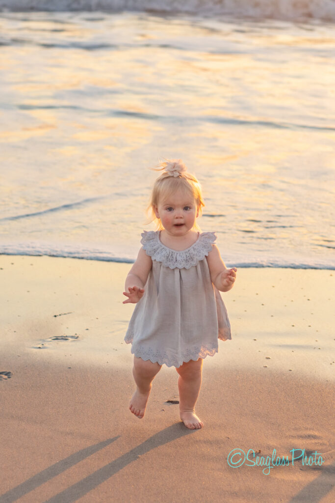 A toddler in a light dress joyfully walks on a sandy beach at sunset. Gentle waves touch the shore in the background, creating a serene atmosphere. Vero Beach Photoshoot