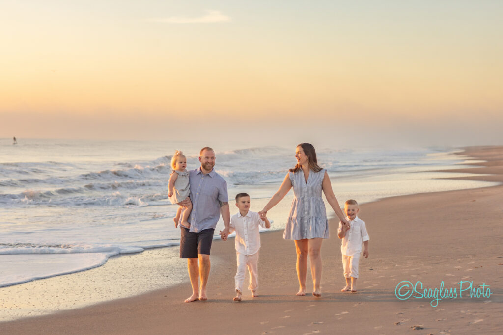 A family of five, including two young boys and a toddler, walks barefoot along a calm beach at sunrise. The parents each hold a childs hand, with gentle waves lapping at the shoreline. The sky is a soft orange and blue. Vero Beach Photoshoot