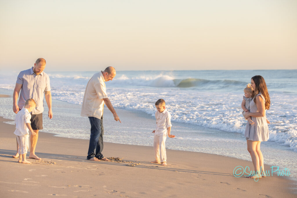 A family enjoys a sunny day at the beach. Two adults and a child stand on the sand near the shore, with one adult pointing to the sand. Another adult holds a baby. Gentle waves roll in under a clear sky. Vero Beach Photoshoot