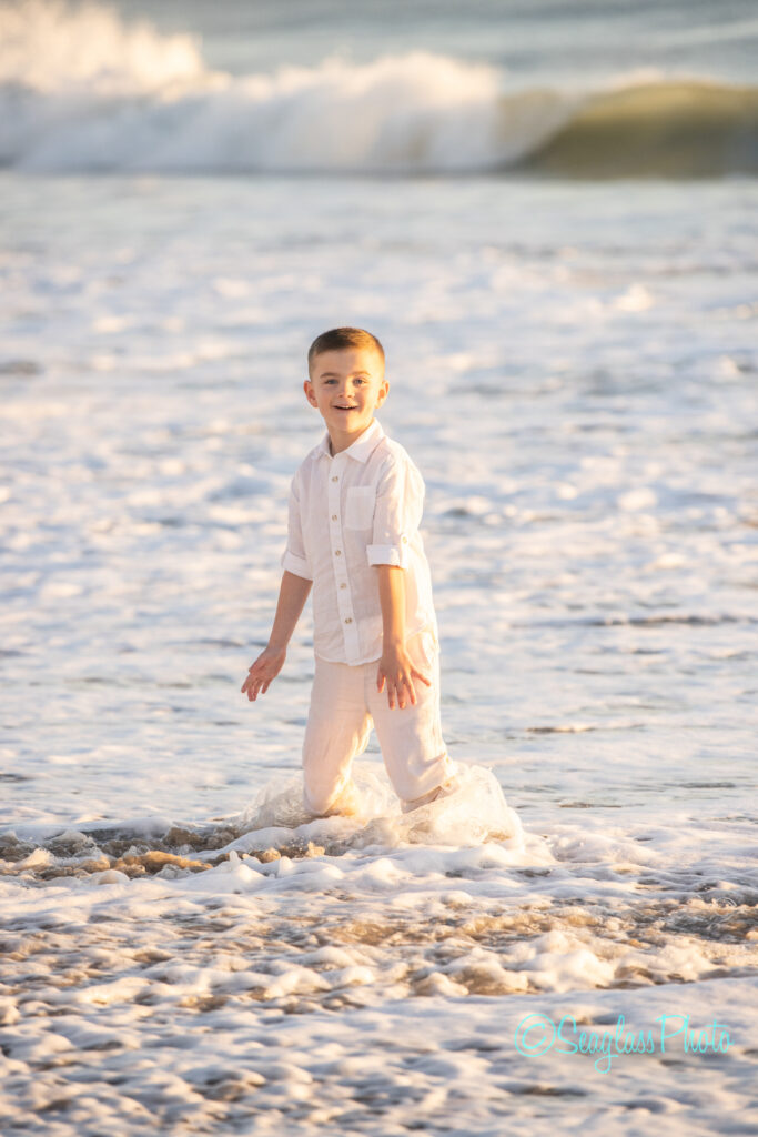 A young boy dressed in white stands at the edge of the ocean, with waves gently crashing around him. The sun casts a warm glow on the water and sand. Vero Beach Photoshoot