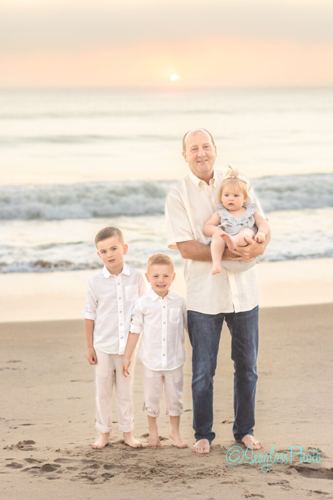 A man stands on the beach with three children. He is holding a toddler, while two young boys stand beside him. They are all wearing light-colored clothing. The ocean and sunset are visible in the background. Vero Beach Photoshoot