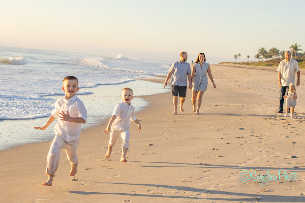 Two young boys joyfully run on a sandy beach, followed by several adults in casual attire. The ocean waves and palm trees are visible in the background under a clear sky. Vero Beach Photoshoot
