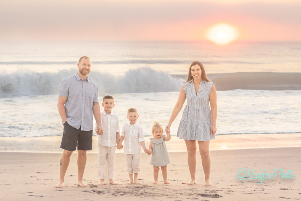 A family of five stands on a beach at sunset, holding hands. The father and mother are on either side, with their three young children between them. The ocean waves are in the background, and the sky is a soft gradient from orange to blue. Vero Beach Photoshoot