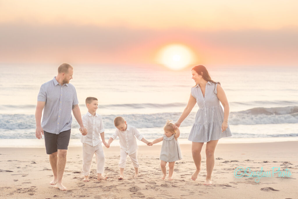 A family of five walks hand in hand on the beach at sunset. The parents are on either side, with two young boys and a toddler girl between them. The sky is colorful, and the sun is low over the ocean. They all wear light summer clothing. Vero Beach Photoshoot