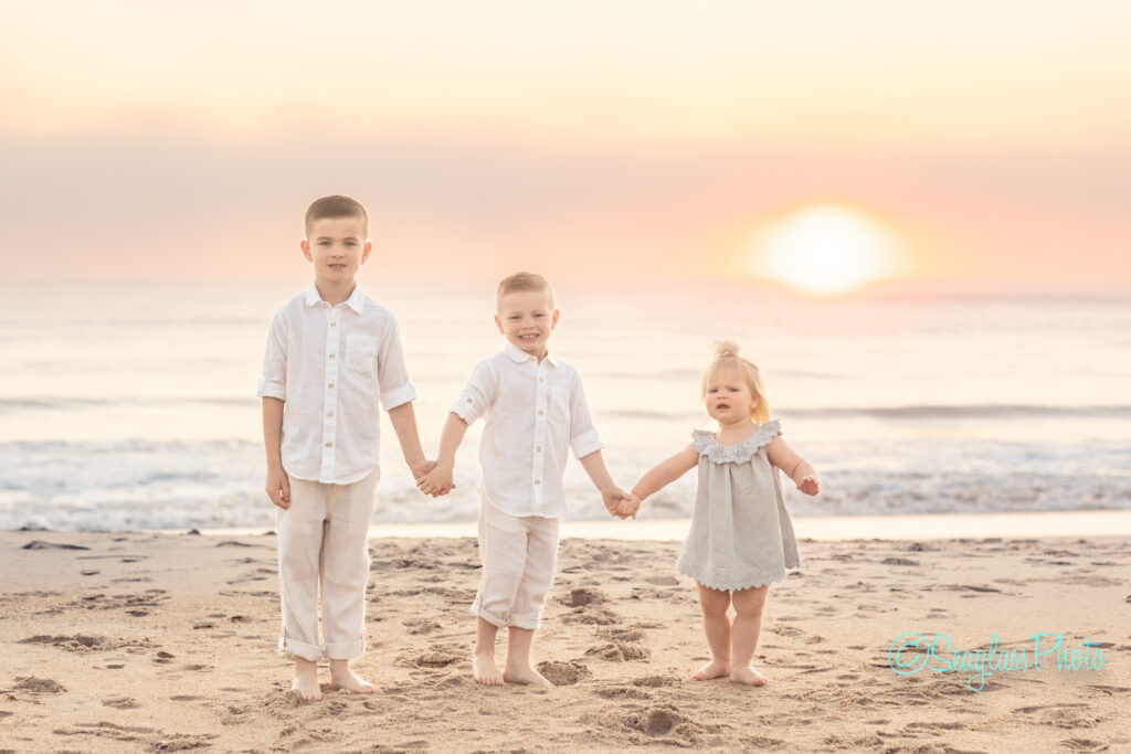 Three young children holding hands on a sandy beach during sunset. They are wearing light-colored clothing, with two boys standing on either side of a younger girl in a dress. The ocean and setting sun are in the background. Vero Beach Photoshoot