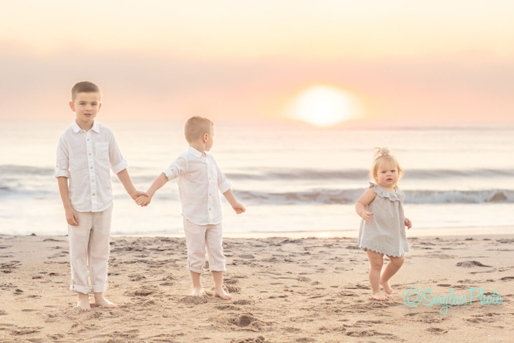 Three young children stand on a beach at sunrise. Two boys hold hands, wearing white shirts and pants. A toddler girl in a light dress stands beside them. The ocean and the sun are in the background. Vero Beach Photoshoot